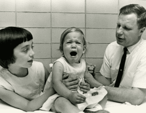 Young child crying in pain from receiving a vaccine with a doctor and her sister holding her