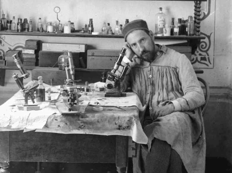 Portrait of Ramón y Cajal, a man seated and resting his head against his hand next to his microscopes