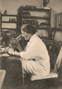 Black and white photo of Lucy Wills, who treated anemia with yeast extract, or marmite. Depicts a white woman with dark hair looking into a microscope. Photo for article on Marmite history and anemia treatment
