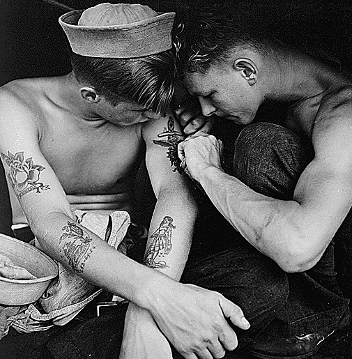 Young man in sailor hat watching other young man create a design on his upper arm. The one being tattooed has designs on his forearms and other upper arm as well.
