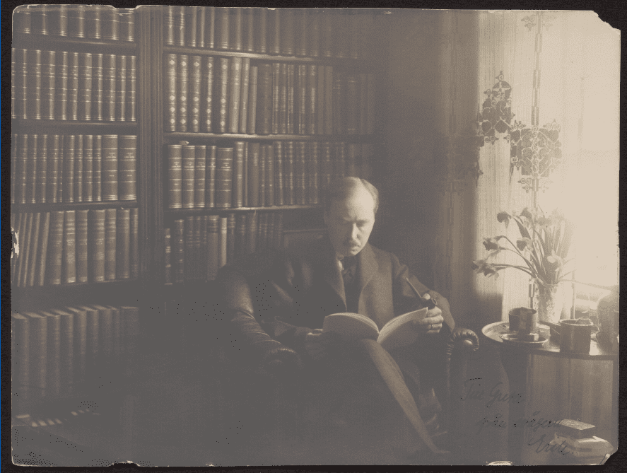 Erik Waller, a man reading, surrounded by books, with an end table with flowers and mugs next to a curtained window. A note in Swedish is written in the bottom right of the photo.
