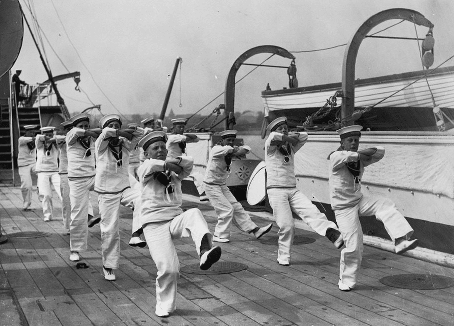 Two lines of six cadets in white with their arms crossed and left legs kicking in the air in example of shipboard dancing in the Navy