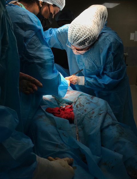 People in scrubs, masks, hairnets, and gloves operating on a patient. The author is wearing glasses and is bent over the patient.